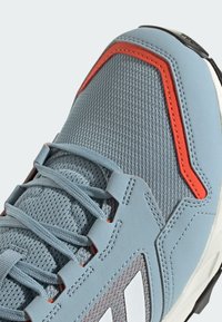 Jasnoniebieski but sportowy z siateczkowym wierzchem, pomarańczowymi akcentami i teksturowanym obszarem sznurowania. Biały logo wyświetlone z boku.
