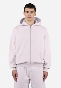 Sudadera con cremallera de color rosa claro, hecha de un tejido suave, que presenta una cremallera frontal, dos bolsillos y puños y dobladillo acanalados. Logo de texto en el pecho.