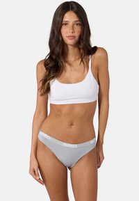 Femme portant un soutien-gorge de sport blanc et un sous-vêtement gris avec le logo PSD sur la ceinture, debout devant un fond uni.