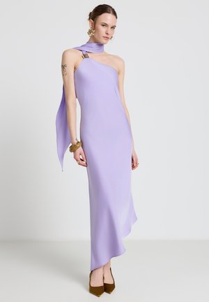 Femme portant une robe lavande sans manches à une épaule avec un détail foulard, des boucles d'oreilles en or, des bracelets et des talons pointus marron, debout sur un sol blanc.