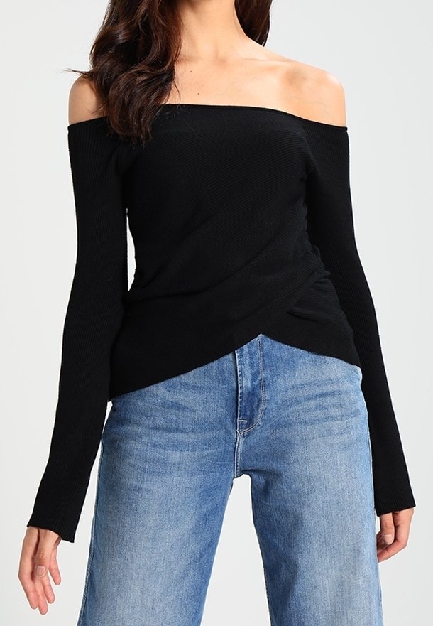 Mujer vestida con una blusa negra de manga larga y escote cuadrado, y vaqueros de tiro alto en azul claro, frente a un fondo liso.
