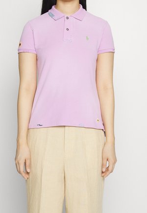 Mujer con una camiseta tipo polo de manga corta color lila claro y pantalones beige, de pie contra un fondo claro y sencillo.