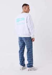 Sudadera blanca con un gráfico en azul claro en la parte posterior, combinada con jeans de mezclilla azul claro que presentan un patrón de texto repetido, y zapatillas grises.