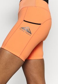 Pantalones cortos deportivos naranjas hechos de material elástico que cuentan con una cintura negra, un bolsillo trasero y un diseño de logotipo reflectante.