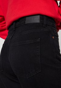 Svart denimjeans med en läderlogotyp, femfickorsdesign och subtila sömmar, tillsammans med en röd sweatshirt.