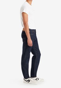 Jeans de mezclilla azul oscuro con un corte clásico, que presentan costuras amarillas en contraste y un parche de cuero marrón en la cintura. Combinados con una camiseta blanca.