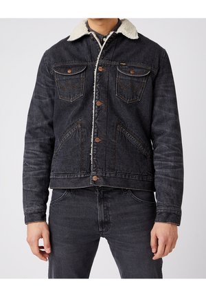 Mann trägt schwarze Jeansjacke mit weißem Fleecefutter und passende schwarze Jeans vor einem schlichten Hintergrund.