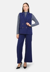 Marinblå ärmlös blazer med dubbelknäppt design, matchad med en mönstrad polotröja och vida byxor, accessoriserad med klackar.