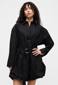 Γυναίκα με σγουρά μαλλιά που φοράει ένα χαλαρό μαύρο φόρεμα με ζώνη, στέκεται με το ένα χέρι στη μέση της μπροστά από ένα απλό φωτεινό φόντο.