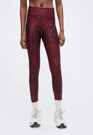 Kvinde iført røde og sorte leggings med høj talje med hvide sokker og hvide sportssko, stående mod en ensartet lys baggrund.