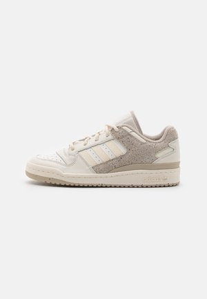 Beżowo-kremowy, niskoprofilowy sneaker Adidas z panelem z zamszu, trzema paskami, perforowanym noskiem i wytłoczonym logo na podeszwie skierowany w prawo.