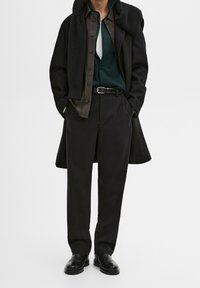Cappotto nero su una camicia verde, sovrapposto a una camicia button-up marrone, cintura nera e pantaloni neri. Tessuti lisci con minimi accenti.