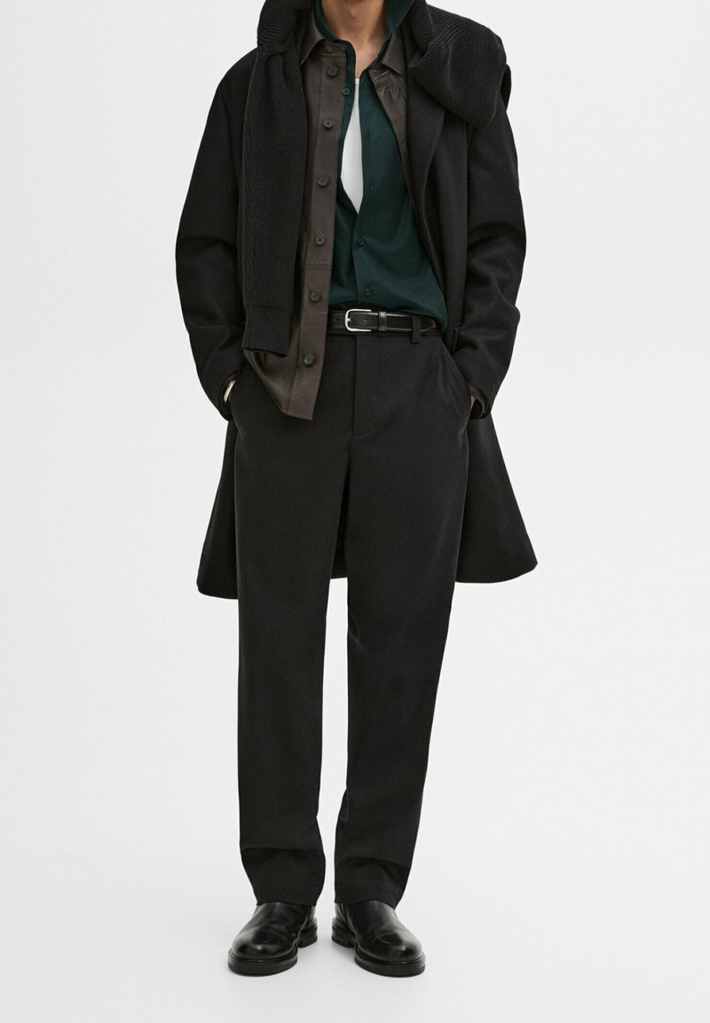 Cappotto nero su una camicia verde, sovrapposto a una camicia button-up marrone, cintura nera e pantaloni neri. Tessuti lisci con minimi accenti.