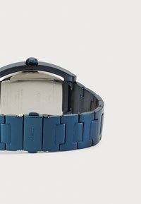 Montre en acier inoxydable bleue avec un boîtier rectangulaire, un bracelet assorti, une finition polie et un logo visible sur le fermoir.