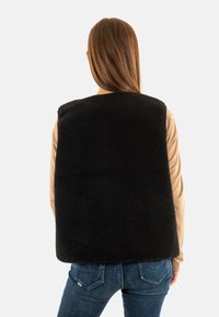 Zwarte fleece vest met een hoekige vorm, tekstuur aan de oppervlakte en zonder sluitingen. Gecombineerd met een lichtbruine langslavige top en blauwe jeans.