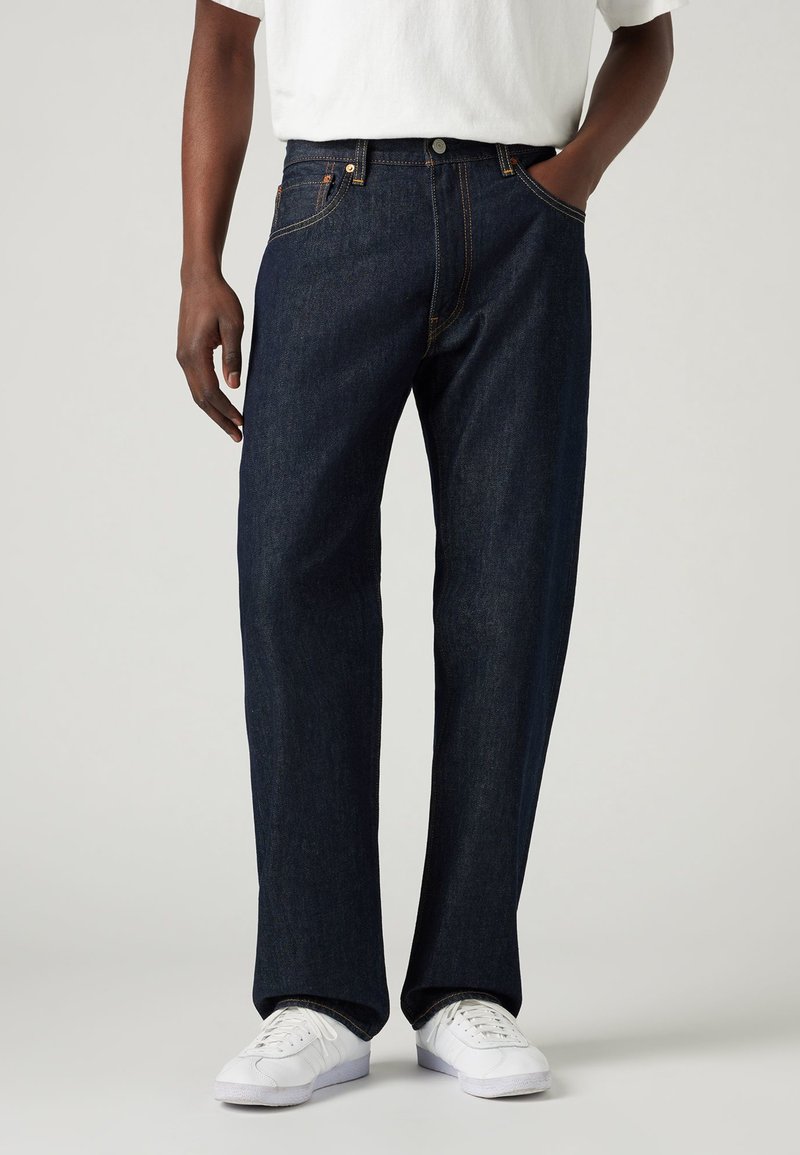 Dunkelblaue Denim-Jeans mit geradem Bein, ausgestattet mit orangen Nähten und Metallbeschlägen. Kombiniert mit weißen Sneakers und einem weißen Hemd.