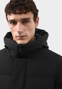 Schwarze Pufferjacke mit hohem, gepolstertem Kragen, Reißverschluss und gestepptem Design. Das Material wirkt glatt und isoliert, geeignet für kaltes Wetter.
