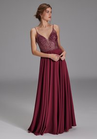 Mujer con un vestido de noche borgoña sin mangas, con escote en V profundo, corpiño con lentejuelas y falda fluida, posando sobre un fondo gris liso.