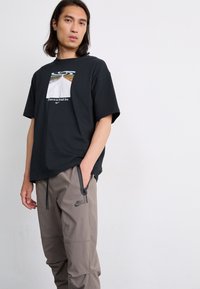 Tricou negru cu mânecă scurtă, având un grafic central și text. Asortat cu pantaloni bej Nike, care prezintă o croială lejeră și o branduire subtilă.