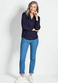 Navyblaues Blusenoberteil mit V-Ausschnitt, langen Ärmeln und subtiler Schimmerung, kombiniert mit hellblauen Jeans mit hochgekrempelten Ärmeln und weißen Sneakers.