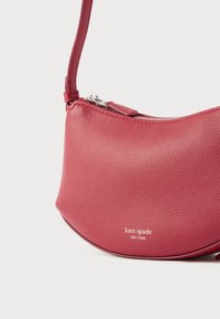 Rdeča usnjena torbica za ramo z teksturirano površino, ukrivljene oblike, z zadrgo in srebrnimi kovinskimi dodatki. Na njej je logo: "kate spade NEW YORK."