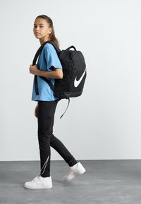 Nike ryggsäck i svart tyg med en framträdande vit swoosh, buren med en blå skjorta och svarta leggings som har vita detaljer.