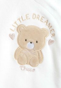Broderad vit tyg med en beige nallebjörn som har en blå nos, en rosett och texten "Little Dreamer" ovanför. Varumärket "Chicco" under.