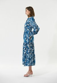 Langes Kleid mit floralen Mustern in Blau und Weiß, mit Puffärmeln, Knöpfen, einem gestuften Rock und seitlichen Taschen. Kombiniert mit weißen Sandalen.
