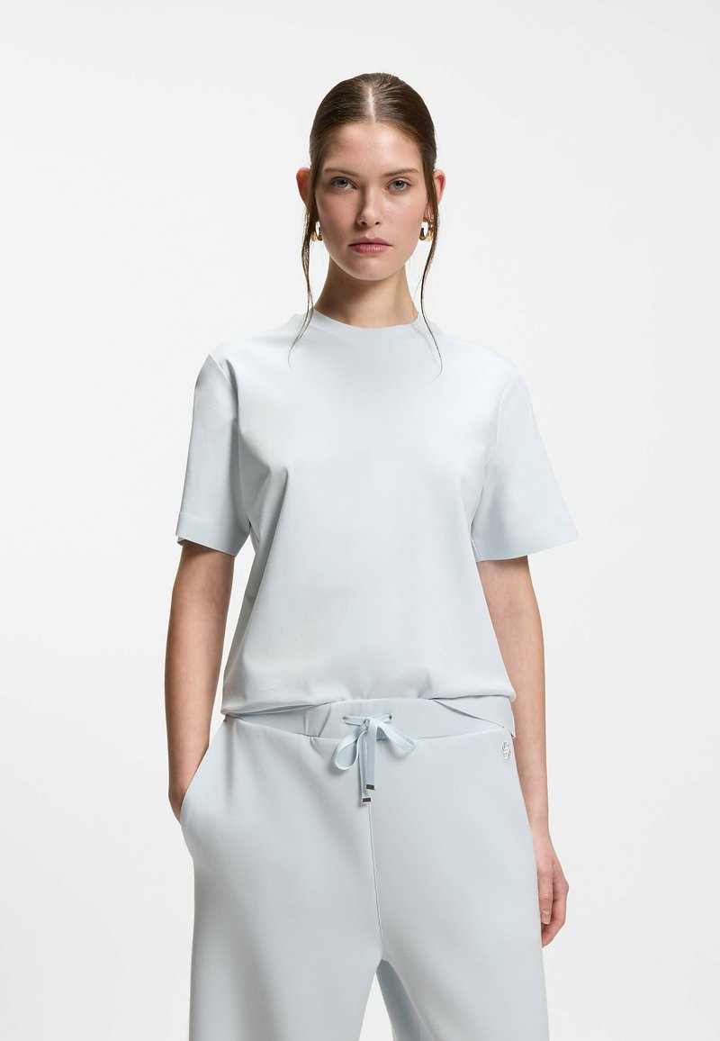 Femme aux cheveux bruns portant un haut à manches courtes blanc et un pantalon assorti avec cordon de serrage, debout devant un fond clair uni, les mains dans les poches.