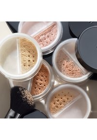 Fem gjennomsiktige beholdere med løst pudder i nyanser av beige, rosa og gul, sammen med en svart sminkebørste, som viser fin tekstur og rom.