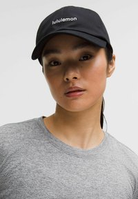 Giovane donna con un cappellino nero Lululemon e una maglietta grigia a girocollo, rivolta in avanti con un'espressione neutra, su uno sfondo semplice.