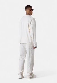 Homme vêtu d'une tenue décontractée entièrement blanche comprenant un haut à manches longues, un pantalon avec poches, des baskets blanches et des lunettes de soleil noires, se tenant de dos.