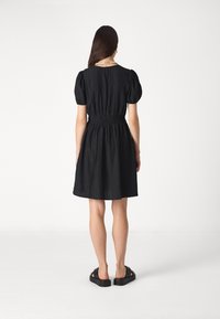 Μαύρο φόρεμα με φουσκωτά μανίκια και σούρα στη μέση, φτιαγμένο από υφασμάτινη υφή. Συνδυασμένο με χοντρές μαύρες σαγιονάρες. Πίσω όψη.