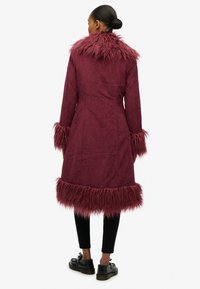 Bordeauxrode jas met een gestructureerde afwerking, voorzien van een pluizige bontkraag en manchetten, en een zoom met franjes. Gedragen over zwarte broek en schoenen.