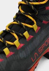 Czarno-czerwony but sportowy z teksturowaną powierzchnią, z żółtymi i czarnymi sznurówkami oraz wyraźnym logo "LA SPORT" w kolorze czerwonym.