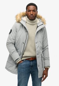 Parka gris acolchado con capucha de piel sintética desmontable, bolsillos frontales y cierre de cremallera, llevado sobre un suéter grueso color crema y jeans.