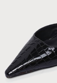 Gros plan d'une chaussure en cuir verni noir à bout pointu avec une texture crocodile et un empeigne basse sur un fond clair uni.