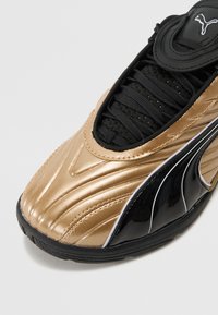 Gull- og svarte sneakers med en glatt, blank overflate, teksturert stoff rundt snøringen og buede designlinjer. Svart gummi såle synlig.
