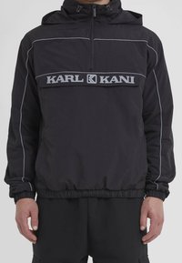 Black hooded jacket with front zipper and "KARL KANI" logo across chest, worn with matching black pants on a standing person.
