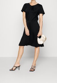 Černé šaty po kolena s krátkými rukávy a nařasenými detaily u výstřihu. Kombinované s černými páskovými podpatky a krémovou saténovou kabelkou.