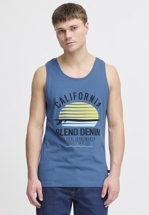 Sinine puuvillane tank top ümmarguse kaelusega, millel on graafiline print päikeseloojangust ning must ja kollane tekst. Koos siniste teksadega.