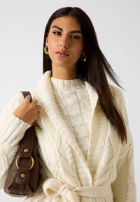 Crème kabelgebreide cardigan met schouderkraag en striktaille, gedragen over een textuurtrui. Houdt een bruine handtas met gouden accenten vast.