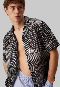 Camicia a maniche corte nera con linee ondulate bianche, dotata di colletto classico e chiusura con bottoni. Abbinata a pantaloni corti viola chiaro con cordoncino.