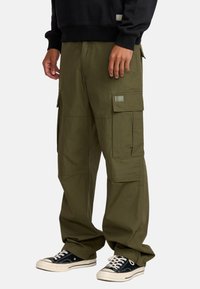 Khaki zielone spodnie cargo z wieloma kieszeniami, luźny krój, mocny materiał i przeszycia. W zestawieniu z czarnymi sneakersami.