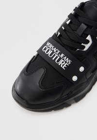 Černá teniska z kůže a síťoviny s výrazným páskem s logem "VERSACE JEANS COUTURE", kulatými bílými detaily a strukturovanou gumovou podrážkou.