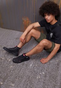 Sneaker alte nere con dettagli rossi e suola texture, abbinate a pantaloni corti verde oliva e una maglietta nera in un contesto di cemento.