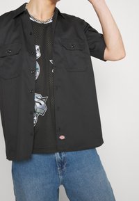 Camisa negra de botones con mangas cortas, que presenta dos bolsillos en el pecho, llevada sobre una camiseta sin mangas de malla gráfica negra, combinada con jeans azules.
