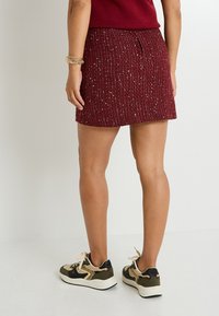 Falda mini de textura roja con acentos de hilo metálico, combinada con zapatillas beige y verde oliva que presentan una mezcla de materiales y patrones.