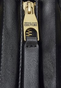 Tirette de fermeture éclair dorée sur cuir noir texturé avec "VERSACE JEANS COUTURE" gravé sur le curseur métallique de la fermeture éclair.