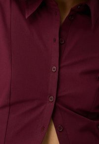 Burgundy skjorta med knappar och spetsig krage, som har bruna knappar och en mjuk textur. Vänster del något öppen, vilket avslöjar underlagret.